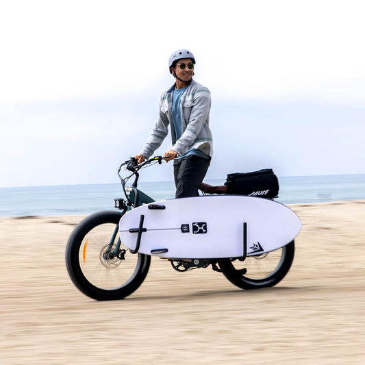 Person riding an electric bike with a surfboard attached on a beach.