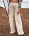 woman wearing beige sweatpants standing on the beach and holding a surfboard.