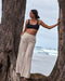 woman wearing a black swim top and beige sweatpants and standing next to a tree