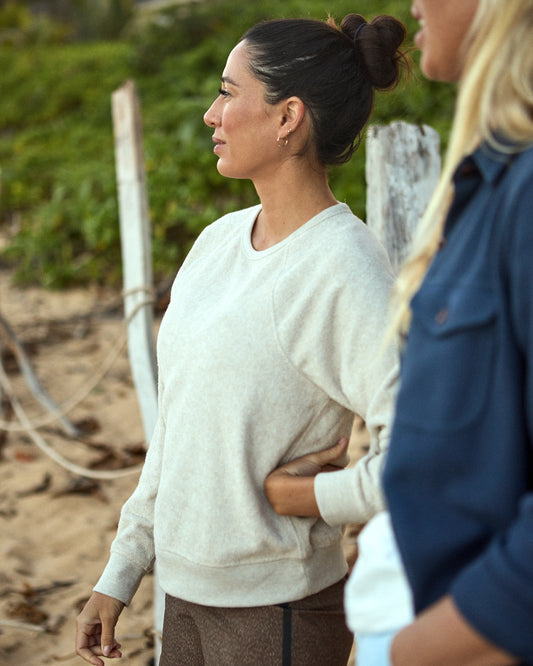 A woman smiling and wearing a cream crewneck sweatshirt, standing on a beach