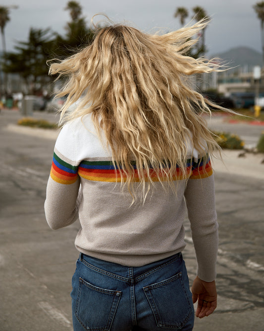 A woman smiling and wearing a beige crewneck sweater with colorful horizontal stripes on the chest area, paired with blue jeans.