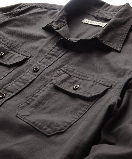 A long-sleeved black shirt with a herringbone texture, featuring a collar, button-up front, and two chest pockets.