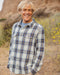 Man wearing a blue, green, and beige plaid shirt in a desert setting with yellow flowers and rocky terrain.