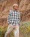Man wearing a blue, green, and beige plaid shirt and beige pants standing in a desert landscape with yellow flowers and rocky cliffs.