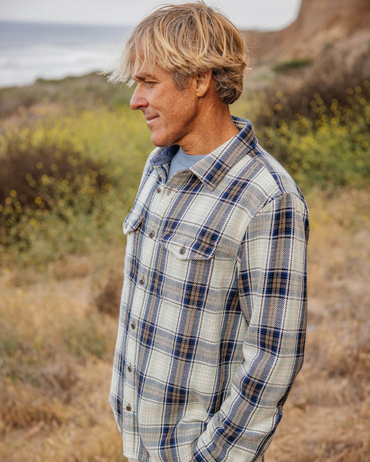 Man wearing a blue, green, and beige plaid shirt standing in a natural setting with grass and wildflowers.