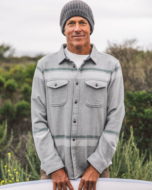 Man wearing a gray flannel shirt and beanie outdoors with greenery in the background