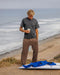Man wearing brown sweats standing on a beach holding a coffee cup with a surfboard and blue jacket nearby.