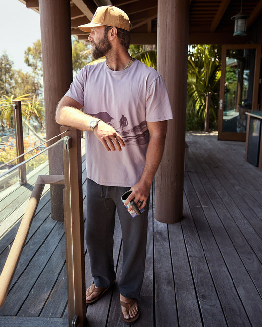 Man standing on a wooden deck holding a can, wearing a cap and casual clothing.