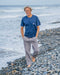 Man standing on a pebbly beach wearing a blue t-shirt and gray pants.