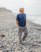 Man standing on a pebbly beach wearing a blue t-shirt and gray pants.