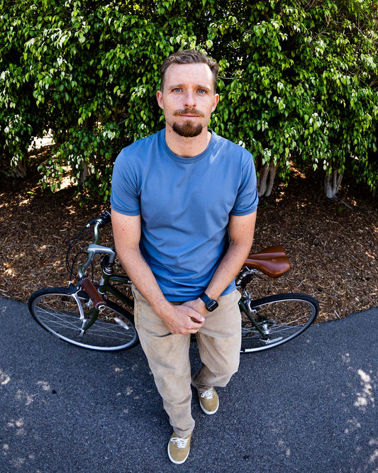 Man in a blue shirt standing next to a bicycle with greenery in the background