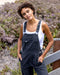 Woman wearing black overalls and a white tank top standing outdoors with purple flowers in the background
