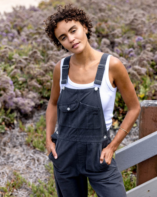 Woman wearing black overalls and a white tank top standing outdoors with purple flowers in the background