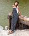Woman wearing gray overalls sitting on a wooden dock by a body of water.