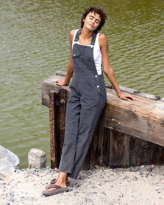Woman wearing gray overalls sitting on a wooden dock by a body of water.