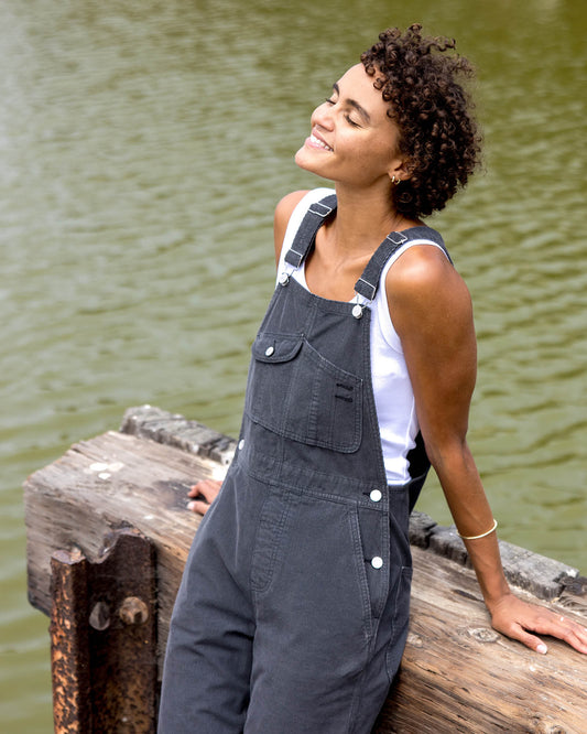Woman wearing black overalls standing on a wooden dock by a body of water.