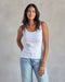 A woman wearing a white ribbed tank top and blue jeans.