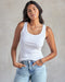 A woman wearing a white ribbed tank top and blue jeans.