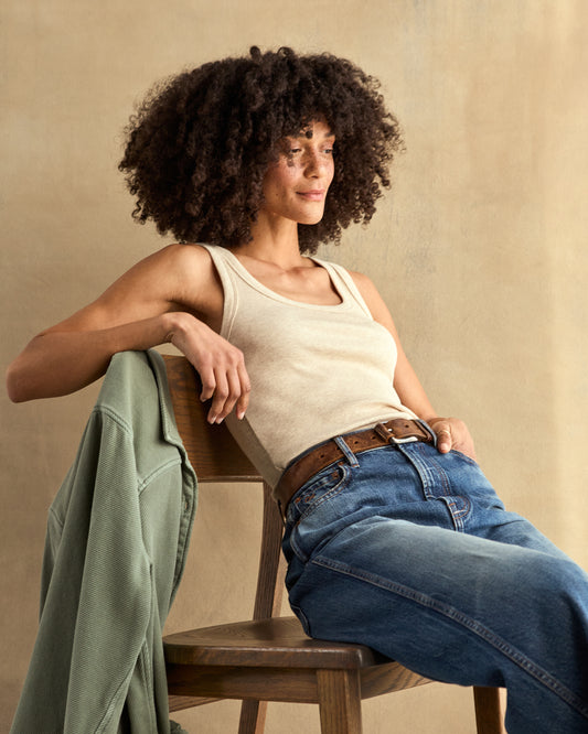 A woman sitting wearing a beige ribbed tank top and blue jeans.