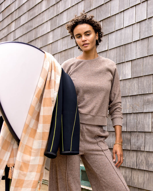 Woman in a beige outfit holding a surfboard against a wooden wall.