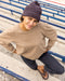 Person wearing a beige sweater and purple-brown beanie sitting on bleachers.
