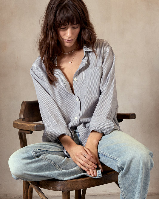 A woman sitting on a wooden chair wearing a black and white pinstriped oversized shirt and light blue jeans.