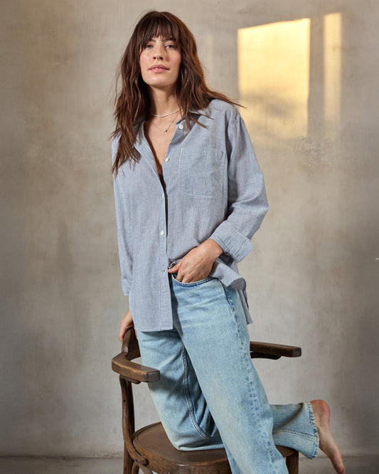 A woman sitting on a wooden chair wearing a black and white pinstriped oversized shirt and light blue jeans.