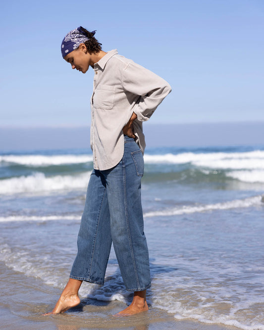 Person wearing a beige shirt and blue jeans standing on a beach with waves in the background