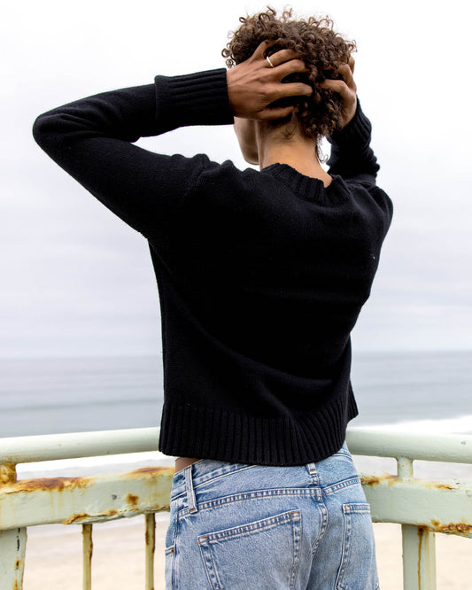 Person wearing a black sweater and light blue jeans standing on a bridge with a cloudy sky.