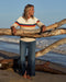 Woman standing on a beach with large driftwood, wearing a colorful sweater and jeans.
