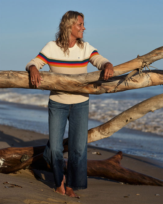 Woman standing on a beach with large driftwood, wearing a colorful sweater and jeans.