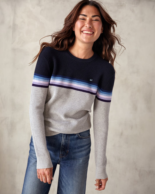 A woman wearing a striped sweater with a crew neckline, where the stripes feature shades of gray, blue, and purple, and the brand logo 'OK' is visible on the chest.