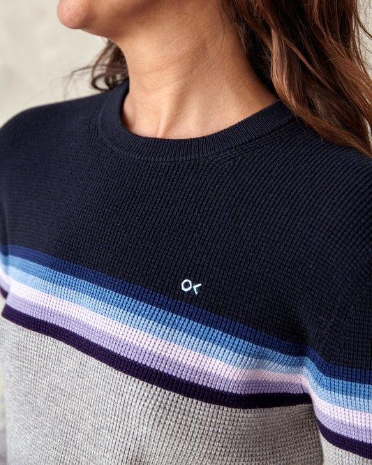 A woman wearing a striped sweater with a crew neckline, where the stripes feature shades of gray, blue, and purple, and the brand logo 'OK' is visible on the chest.