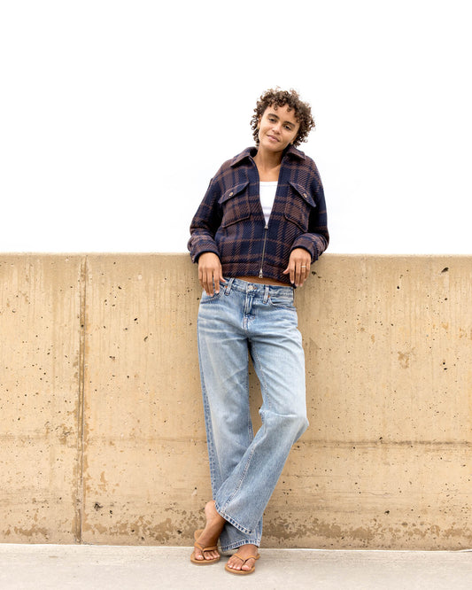 Person wearing a plaid jacket and jeans leaning against a concrete wall.