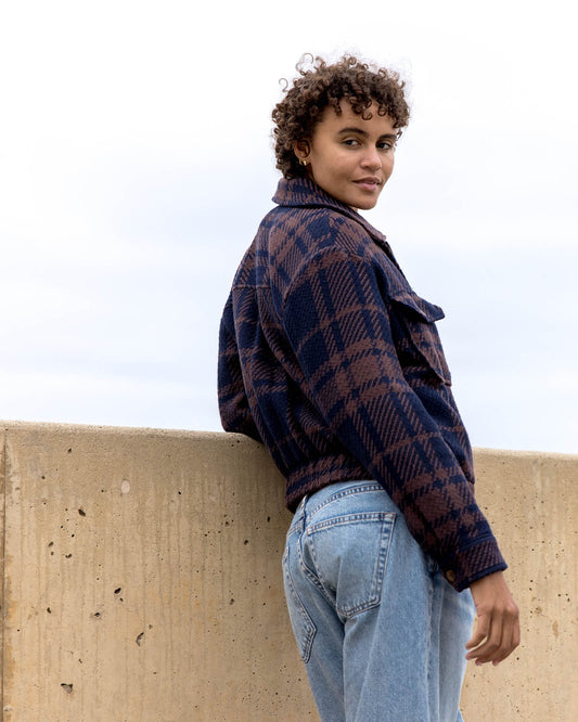 Person wearing a plaid jacket and jeans leaning against a concrete wall with a light background