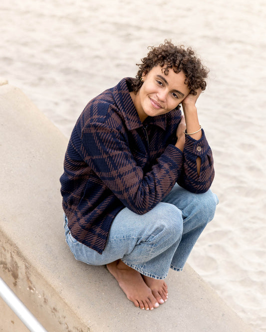 Person sitting on a sandy beach wearing a plaid jacket and jeans.