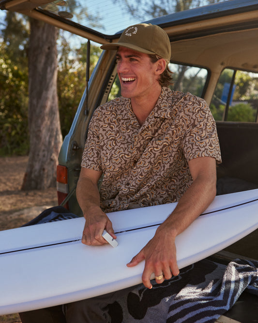 Man wearing brown patterned shirt sitting on the trunk of car waxing a surfboard 
