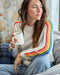 Woman sitting on a couch wearing a sweater with rainbow sleeves, holding a mug.