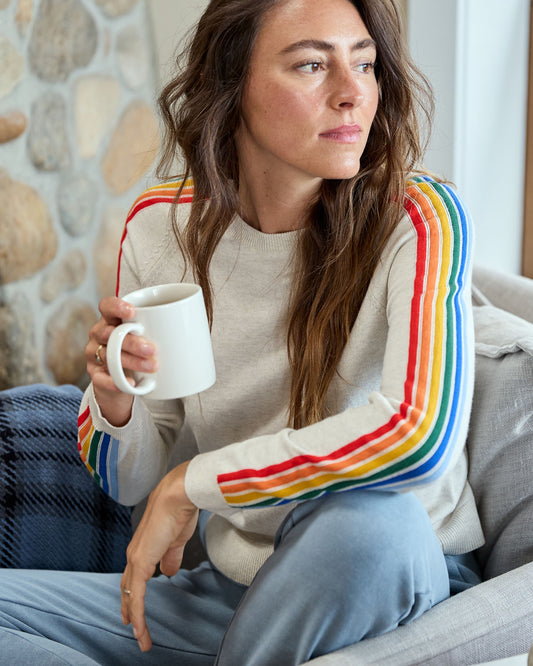 Woman sitting on a couch wearing a sweater with rainbow sleeves, holding a mug.