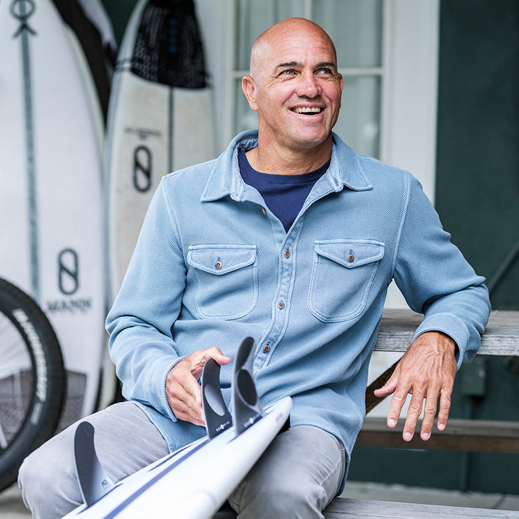 Man sitting with a surfboard, wearing a light blue shirt and gray pants, with surfboards in the background.