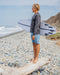 Man holding a surfboard on a pebbly beach with ocean and mountains in the background