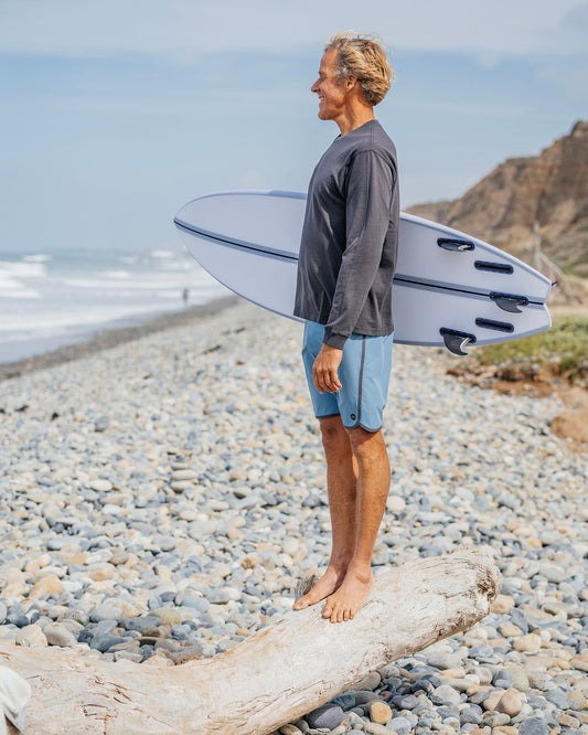 Man holding a surfboard on a pebbly beach with ocean and mountains in the background