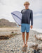 Man holding a surfboard on a pebbly beach with ocean and sky in the background