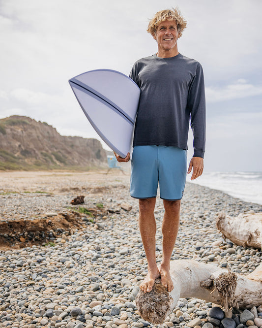Man holding a surfboard on a pebbly beach with ocean and sky in the background