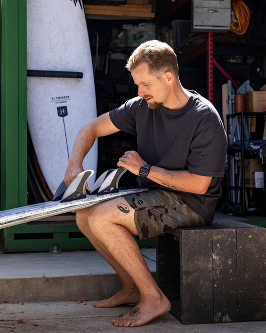 Man sitting on a block in a garage, working on a surfboard.