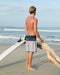 Man standing on a beach with a surfboard, looking out at the ocean.
