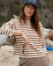 Woman wearing a striped shirt and cap leaning against driftwood on a beach.