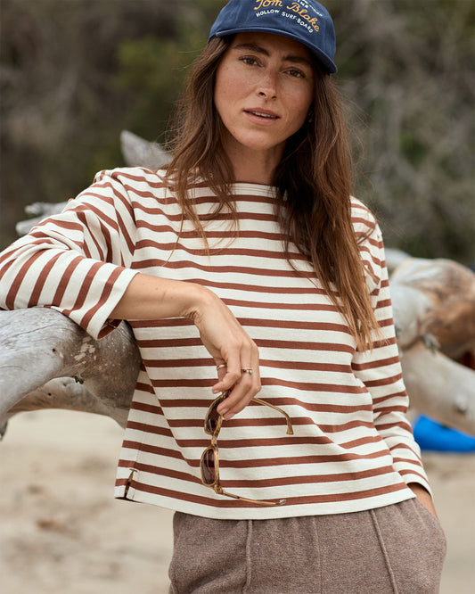 Woman wearing a striped shirt and cap leaning against driftwood on a beach.