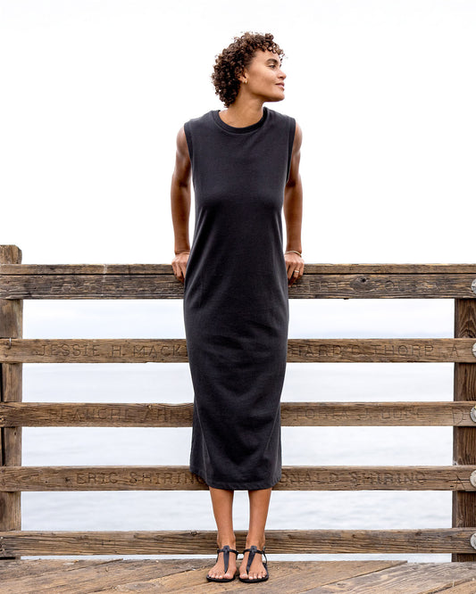 Woman wearing a black sleeveless dress standing against a wooden fence.