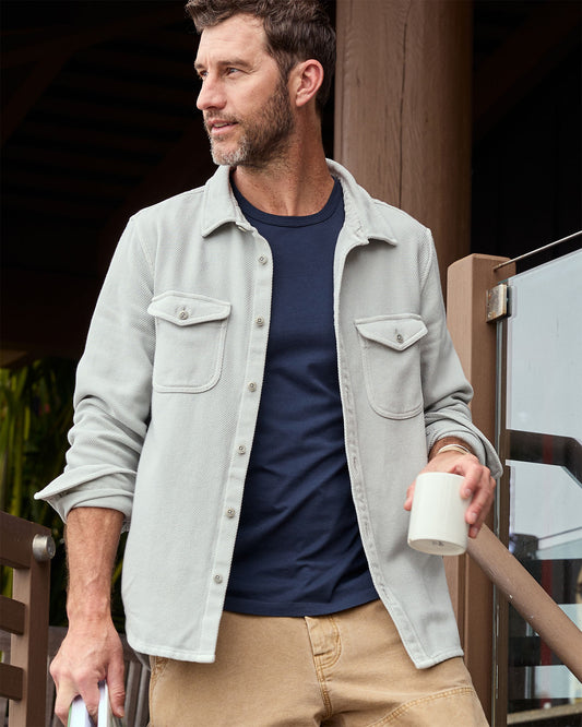 Man wearing a light gray shirt over a navy shirt, holding a white mug, standing outdoors.